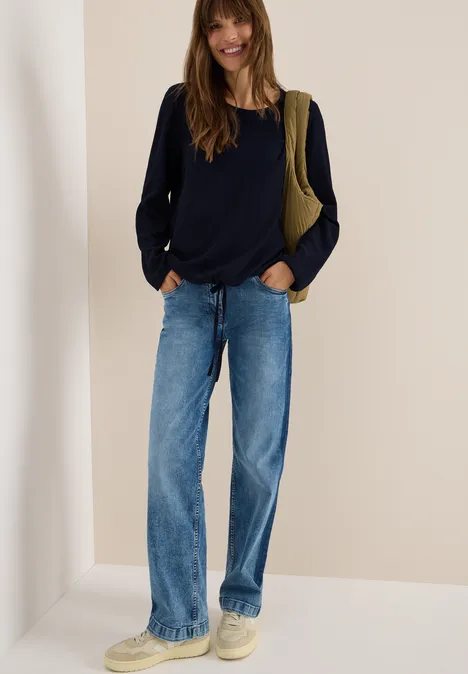 Frau mit dunkelblauem Pullover, Jeans und beiger Tasche steht lächelnd vor hellem Hintergrund.